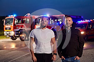 Group of firefighters, dressed in civilian clothing, stand in front of fire trucks during the night, showcasing a moment