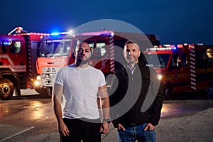 Group of firefighters, dressed in civilian clothing, stand in front of fire trucks during the night, showcasing a moment