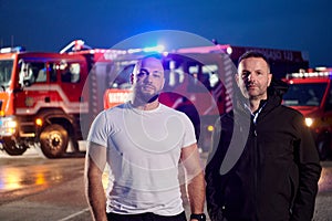 Group of firefighters, dressed in civilian clothing, stand in front of fire trucks during the night, showcasing a moment