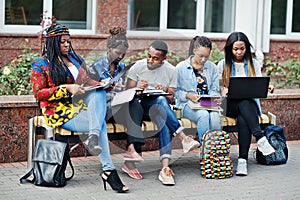 Group of five african college students spending time together on campus at university yard. Black afro friends studying at bench