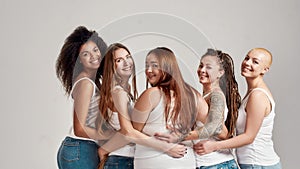 Group of five beautiful diverse young women wearing white shirt and denim jeans putting arms around each other, looking