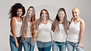 Group of five beautiful diverse young women wearing white shirt and denim jeans smiling at camera while posing together