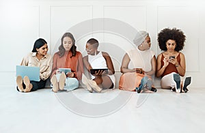 Group of five businesswomen sitting on the floor against a wall in an office and using tech. Businesspeople and