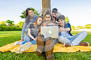 Group of five happy young diverse multiracial gen z people sitting on the grass in a city park field using laptop to surfing net.