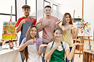 Group of five hispanic artists at art studio amazed and smiling to the camera while presenting with hand and pointing with finger