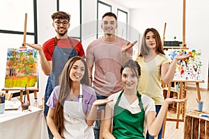 Group of five hispanic artists at art studio smiling cheerful presenting and pointing with palm of hand looking at the camera
