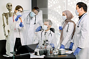 Group of five multiethnic laboratory scientists in lab coats, masks and gloves, working at lab with test tubes