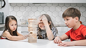 Group of focused children playing board game Jenga, building tower made of wooden blocks beams. Worried girl cover eyes.