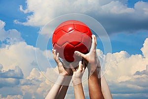 Group of football fans holding red soccer ball in blue cloudy sky. Kid s and adult s hands together hold ball