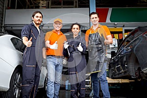 Group of four car service technician men and woman giving thumbs up to camera, people working together at vehicle repair garage
