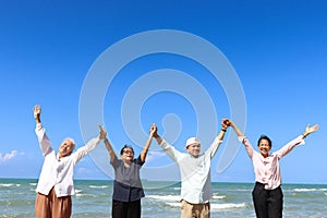 Group of four cheerful elderly old senior friends travel outdoor together, holding hand in line and raising up on beach blue sky,