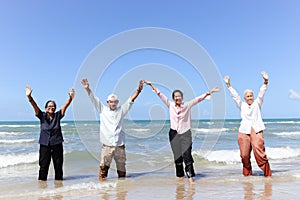 Group of four cheerful elderly old senior friends travel outdoor together, raising hands while running on beach, have fun and