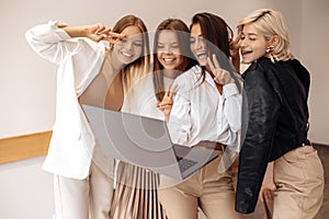 Group of four fashionable young women friends chatting on computer showing peace gesture at the cafe indoors.