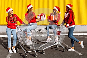 Group of four happy young women in christmas sweaters and santa claus hats with christmas gifts on carts near shopping mall