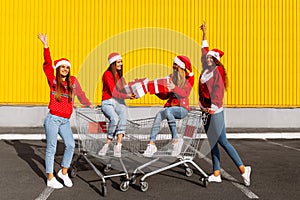 Group of four happy young women in christmas sweaters and santa claus hats with christmas gifts on carts near shopping mall