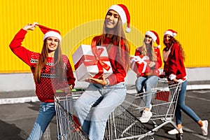 Group of four happy young women in christmas sweaters and santa claus hats with christmas gifts on carts near shopping mall