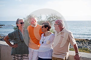 Group of four mature people having fun and talking together at the beach - pensioners seniors smiling and laughing with the sea at