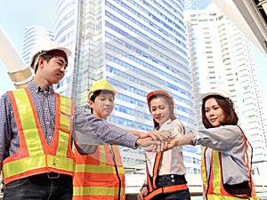 Group of four young Asian engineers wearing safety vest and helmet, putting hands join together at outside office in downtown city