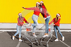 Group of four young women in Christmas sweaters and Santa Claus hats having fun on carts near a shopping center in the city