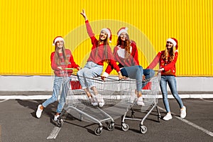 Group of four young women in Christmas sweaters and Santa Claus hats having fun on carts near a shopping center in the city