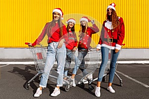 Group of four young women in Christmas sweaters and Santa Claus hats having fun on carts near a shopping center in the city