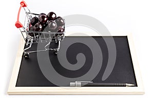 Group of fresh red cherries in a small shopping cart next to a pen on a black chalkboard