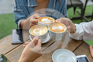 Group of friends cheers with latae cup in cafe bar with phone on table sitting outdoor at cafe - Young girl group having fun photo