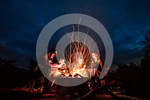 Group of friends chill and warm near a bonfire in camping during a hiking trip.