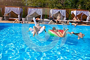 Group of friends chilling in private villa swimming pool lie in the sun on inflatable flamingo, swan, mattress. Young