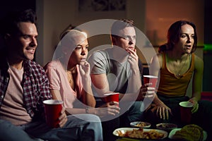 Group Of Friends With Drinks Sitting On Sofa At Home Watching Horror Movie Together