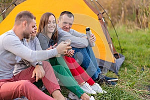 A group of friends is enjoying a warming drink from a thermos, on a cool evening by a fire in the forest. Fun time camping with