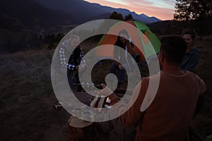 Group of friends gathering around bonfire at camping site in evening