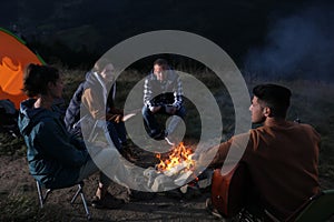 Group of friends gathering around bonfire at camping site in evening