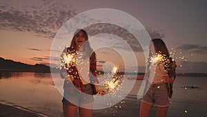 A group of friends girls and men dance on the beach with sparklers in slow motion at sunset. Celebrate new year on the