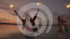 A group of friends girls and men dance on the beach with sparklers in slow motion at sunset. Celebrate new year on the