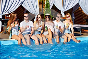 Group of friends having fun at poolside party clinking glasses with fresh cocktails sitting by swimming pool on sunny