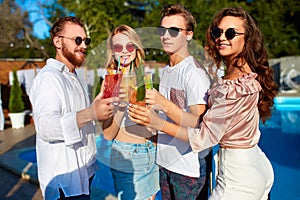 Group of friends having fun at poolside summer party, clinking glasses with colorful summer cocktails outdoors near