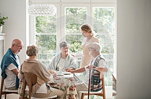 Group of friends with helpful carer sitting together at the table at nursing home dining room