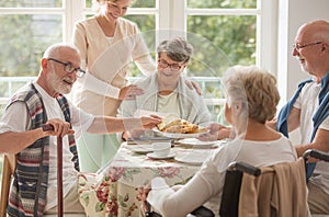 Group of friends with helpful carer sitting together at the table at nursing home dining room and eating cake