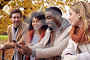 Group Of Friends Outdoors Wearing Coats And Scarves Looking At Photos On Phone In Autumn Park