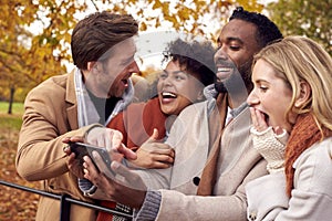 Group Of Friends Outdoors Wearing Coats And Scarves Looking At Photos On Phone In Autumn Park