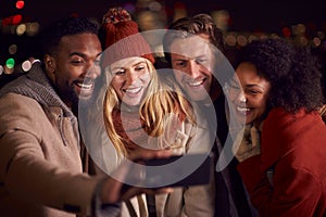 Group Of Friends Outdoors Wearing Coats And Scarves Posing For Selfie On Phone Against City Lights