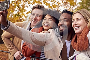 Group Of Friends Outdoors Wearing Coats And Scarves Posing For Selfie On Phone In Autumn Park