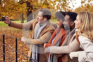 Group Of Friends Outdoors Wearing Coats And Scarves Posing For Selfie On Phone In Autumn Park