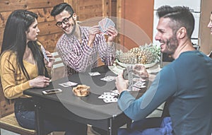 Group of friends playing cards sitting at table inside holidays camping house bungalow - Young happy people having fun laughing