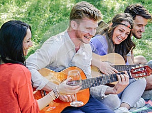 Group of friends playing guitars and laughing while drinking red wine sitting on grass in a park outdoor