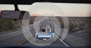 Group Of Friends On Road Trip Driving Classic Convertible Car Viewed Through Windshield Of Following Vehicle