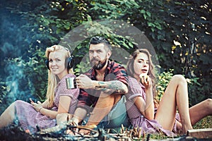 Group of friends sitting next to campfire. Girls leaning on their male friend. Blond woman listening to music while