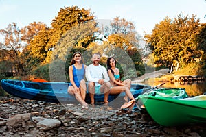 A group of friends are sitting posing on kayaks on the river bank. Active recreation of young people in nature. Concept