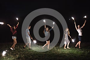 A group of friends with sparklers standing outdoors at dusk.